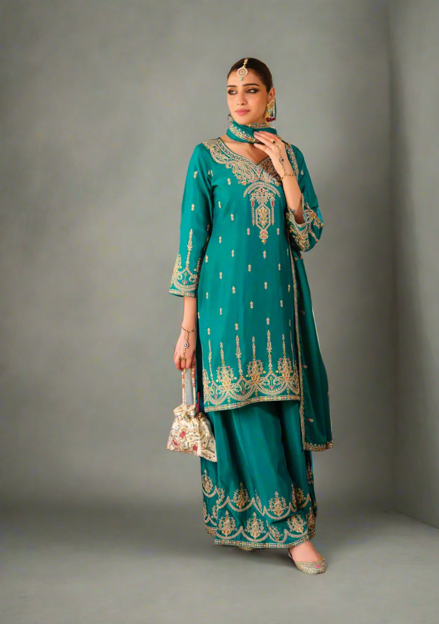 Woman in teal embroidered outfit standing in a decorated room with a table set for dinner.